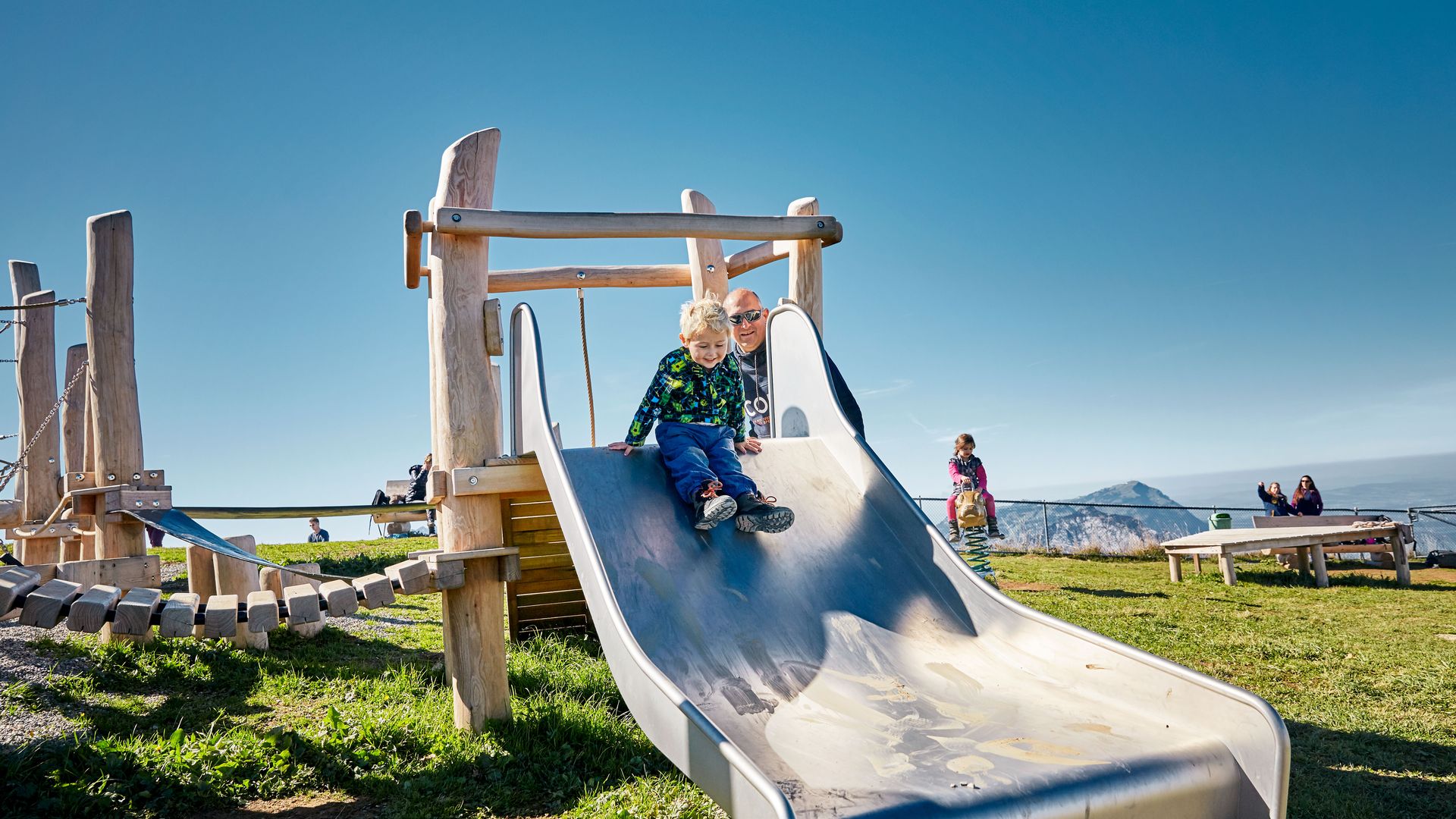 Child sliding down a slide on a playground in an alpine setting.